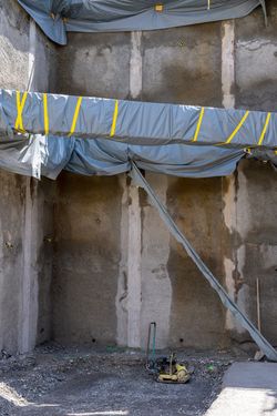 Baustelle mit grauen Betonwänden, Schutzplanen, gelben Gurten und einer gelben Rüttelplatte auf Schotterboden.