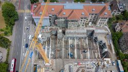 Luftaufnahme einer Baustelle mit großer Baugrube, gelbem Kran, Arbeitern, angrenzendem Gebäude und Straße.