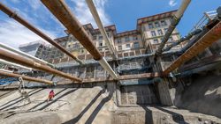 Baustelle mit tiefer Baugrube, horizontalen Rohren, einem Arbeiter und einem alten Gebäude.