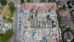 Luftaufnahme einer großen städtischen Baustelle mit gelbem Turmkran, tiefer Baugrube, Baufahrzeugen, einer Straße und Gebäuden.