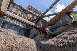 Tiefe Baugrube mit umfassender Metallabstützung und umwickelten Stützen neben einem verzierten Altbau unter blauem Himmel.