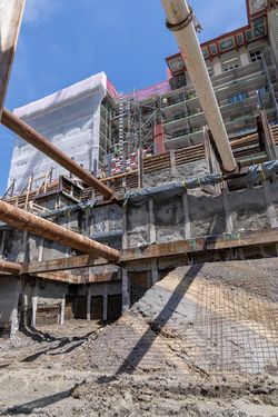 Eine tiefe urbane Baugrube mit großen rostigen Stützrohren, Betonwänden, Holzverbau und einem eingerüsteten Altbau.
