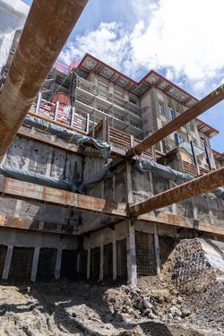 Baustelle mit tiefer Baugrube, großen rostigen Rohren, Betonwänden und einem eingerüsteten historischen Gebäude bei blauem Himmel.