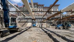 Baustelle mit tiefer Baugrube, mehreren Baggern, Arbeitern und temporären Stützstrukturen unter blauem Himmel.