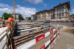 Bauarbeiter betrachtet eine tiefe urbane Baugrube mit Holzschalung, Metallrohren, einem Kran und Gebäuden.